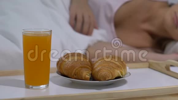 提供果汁和新鲜牛角面包在靠近睡觉女士的托盘中早餐在床上浪漫视频的预览图