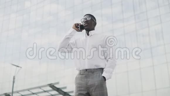一位戴眼镜的非裔美国商人站在办公室附近用手机说话视频的预览图