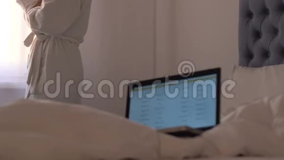 女士穿着浴袍喝着果汁看着窗户笔记本电脑在床上自由职业视频的预览图