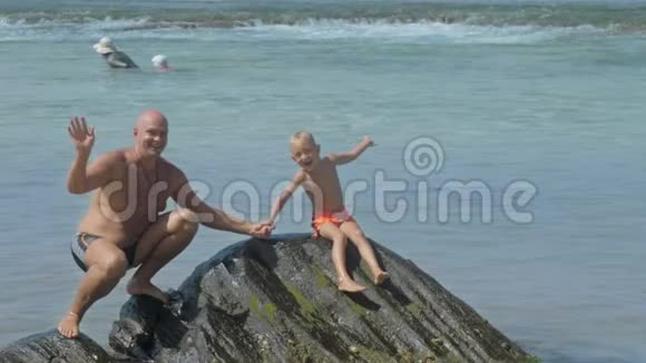 爸爸和男孩坐在石头上挥动双手面对蔚蓝的海洋视频的预览图