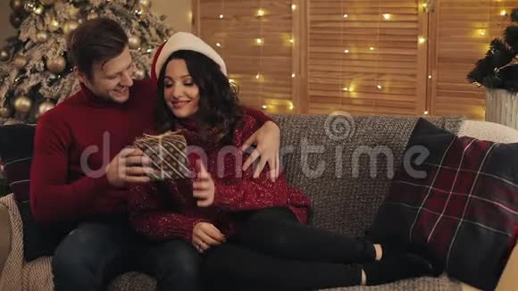 年轻漂亮的男人把他可爱的妻子礼物盒放在圣诞树附近的沙发上亮着亮灯视频的预览图