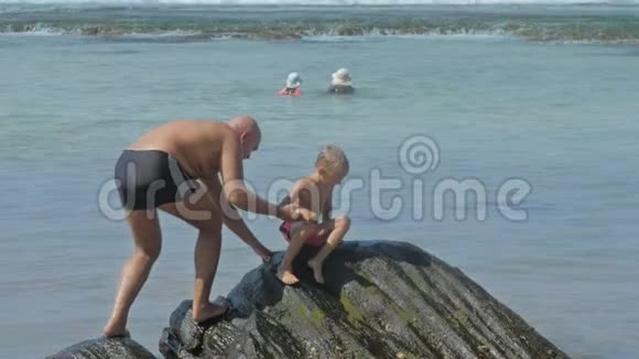 在度假胜地人们帮助小儿子在平静的海洋中攀爬岩石视频的预览图