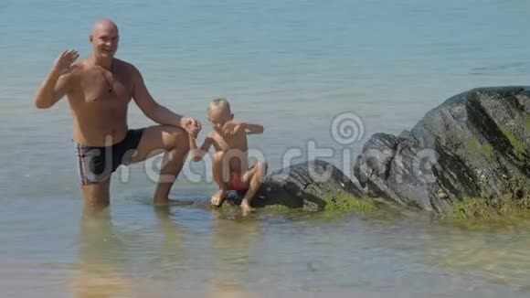 父亲和可爱的儿子在浅海的石头上挥手视频的预览图