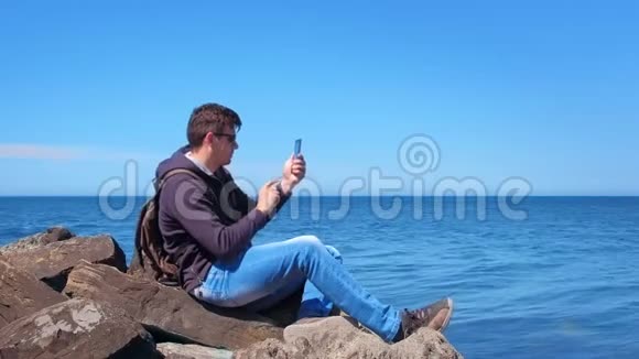 年轻人在智能手机上拍照的游客坐在海边的巨石上视频的预览图
