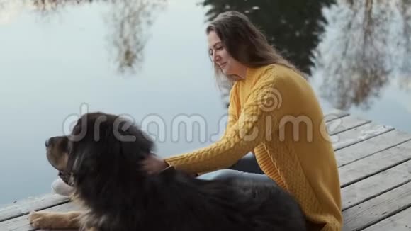 一个穿黄色毛衣的年轻女人坐在码头上抚摸着一只大狗水中树木的倒影视频的预览图