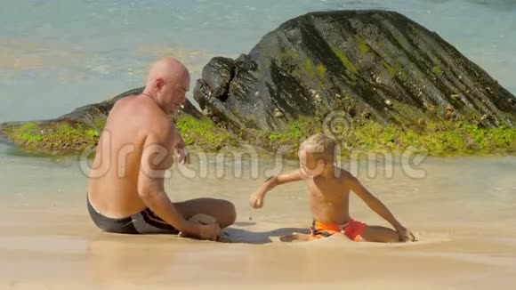 爸爸和可爱的儿子把湿沙扔在沙滩上玩水视频的预览图