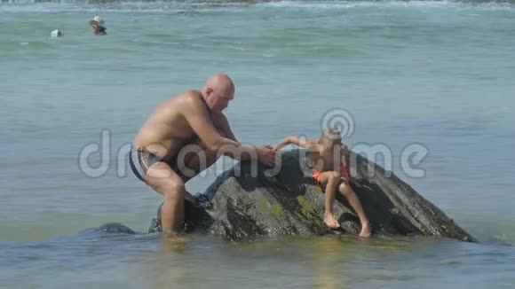 父亲帮助孩子在温暖的海水中从潮湿的岩石上下来视频的预览图