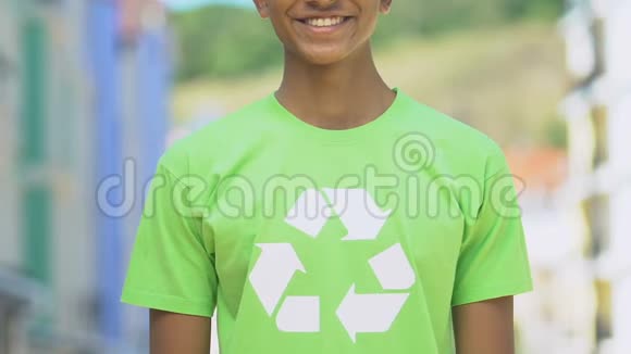穿着带回收标志的衬衫的男孩微笑着进入相机推广可重复使用的产品视频的预览图