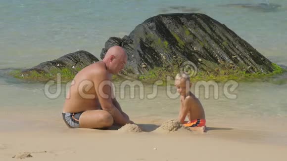 爸爸和可爱的男孩在海滩上用湿沙建造视频的预览图