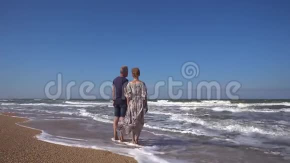 一个身材苗条的男人和一个穿着长裙的女人站在沙滩上迎风破浪视频的预览图