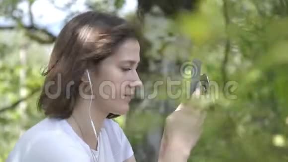 年轻女子的肖像用电话和耳机听音乐欣赏声音公园森林背景年轻女子视频的预览图