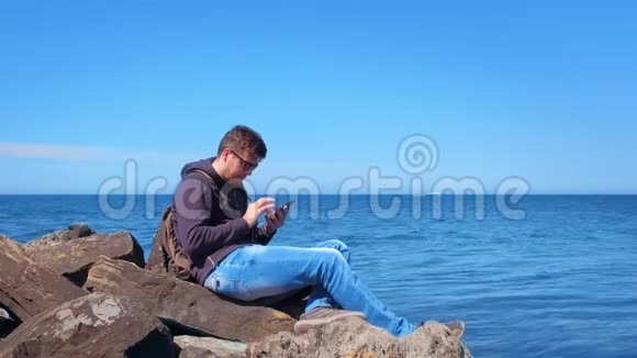 年轻的游客浏览智能手机坐在海边的大石头上视频的预览图