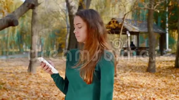 秋天在黄树的背景下漂亮的黑发女人在智能手机上行走和滚动内容视频的预览图