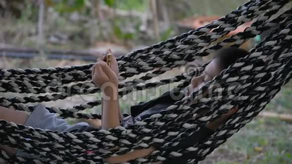 亚洲男孩拿着电话在吊床秋千上休息由花园里的布料制成视频的预览图