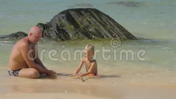 孩子和父亲坐在蔚蓝的海水中坐在岩石上视频的预览图
