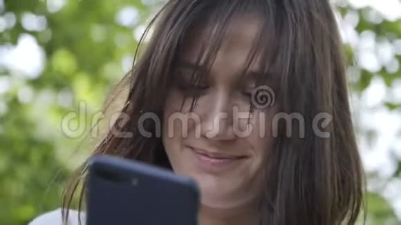 微笑女孩在公园或森林里打短信的肖像女人在电话里聊天通过网络阅读新闻视频的预览图