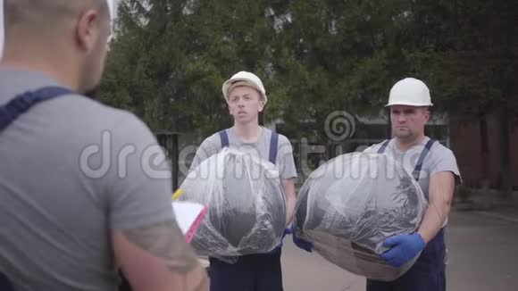 两个戴着白色头盔和蓝色制服的白种人割草机拿着包裹听着他们的首领工人视频的预览图