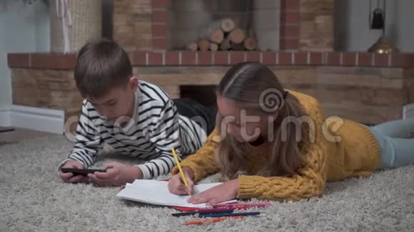 特写白种人可爱的女孩用蜡笔着色看着她的哥哥在玩智能手机游戏兄弟姐妹视频的预览图