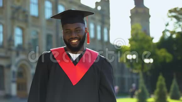 毕业礼服上的黑人男性在大学背景上微笑着视频的预览图