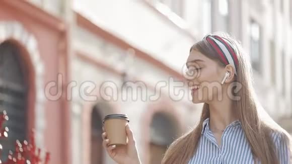 带着时髦头巾和带条纹衬衫的年轻漂亮女孩戴着耳机喝着咖啡的笑容视频的预览图