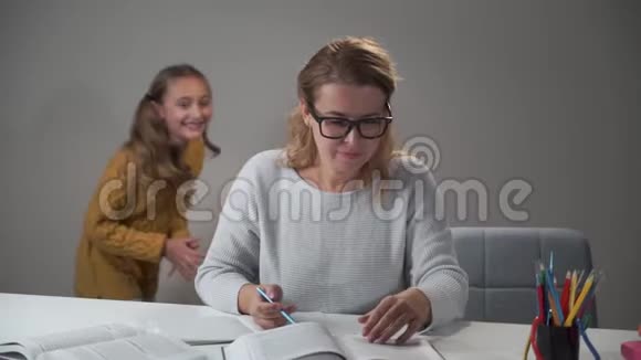 疲惫的白种人女人戴着眼镜坐在桌子旁读着两个快乐的孩子跑来跑去美丽美丽视频的预览图