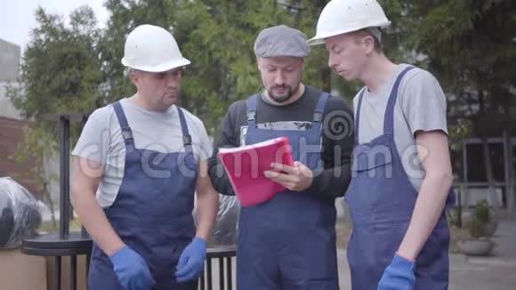 两个戴着头盔和蓝色制服的成年白人搬运工和他们的首领站在一起三名工人正在阅读运单或视频的预览图