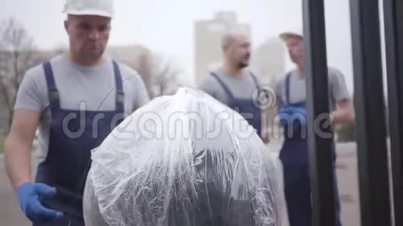戴着白色头盔蓝色手套和制服的严肃的高加索搬运工拿着包裹和走路肌肉男送东西视频的预览图