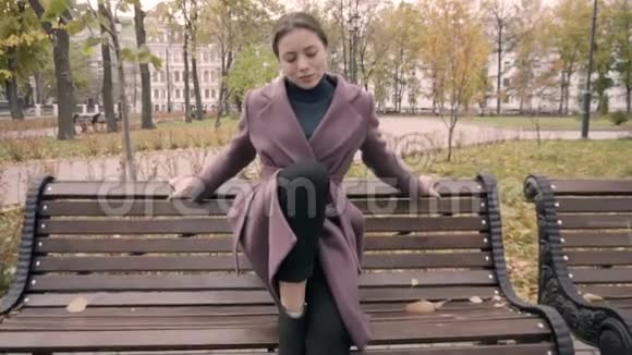 穿大衣的女孩把一只脚放在公园的长凳上视频的预览图