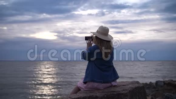 一个年轻的女人坐在石头上在电话上拍照女孩拍摄日落的天空4K慢慢视频的预览图