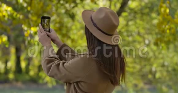 穿着棕色帽子和外套的漂亮黑种人女孩在她的智能手机上拍摄秋天公园的照片愉快的欧洲视频的预览图
