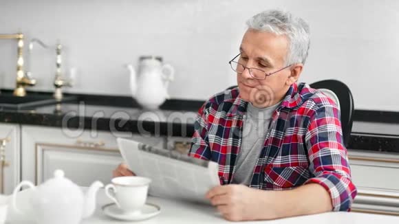 在厨房吃早餐时戴眼镜看报纸的中年家庭男人视频的预览图