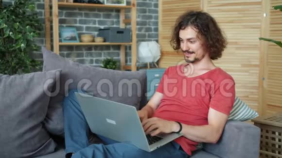 微笑的家伙在家里的沙发上用笔记本电脑打字微笑在网上工作视频的预览图