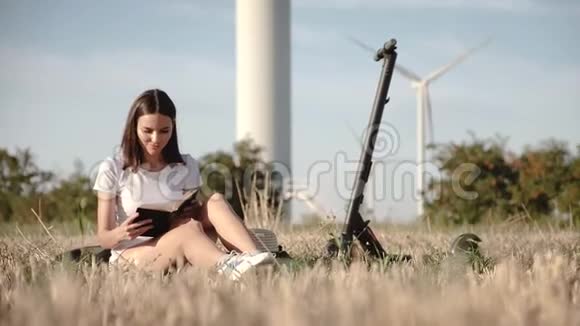 一个迷人的女孩坐在电动滑板车旁边背景是风力发电站和树叶穿过一个电动滑板车视频的预览图