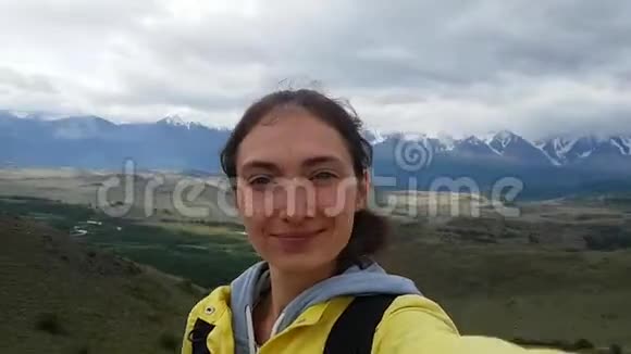 一个身穿鲜黄色夹克的黑发女孩在美丽的山野背景下自拍对着镜头挥手视频的预览图