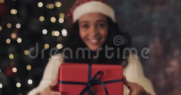 合上戴着桑塔斯帽子的年轻微笑的穆拉托女孩把带蓝色丝带的红色礼物盒拿回来视频的预览图