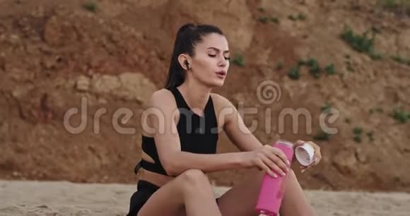 镜头前的特写镜头口渴的女士在经过艰苦的锻炼后从海滩上的一个粉红色的运动瓶里喝水视频的预览图