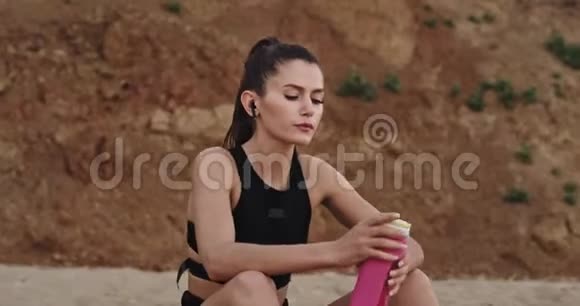 疲惫的女士坐在沙滩上喝着口渴的水早上她在海边努力锻炼后视频的预览图