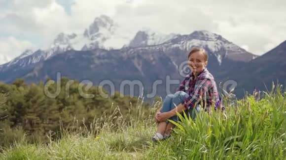 阳光明媚的一天穿着格子衬衫的微笑的女人坐在一片绿色的草地上阿尔卑斯山视频的预览图
