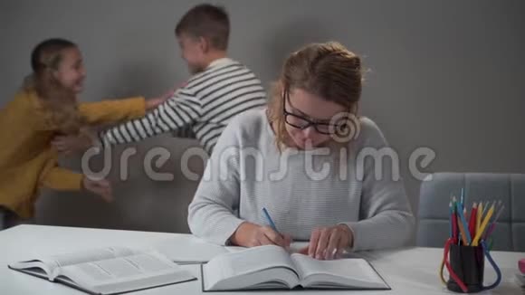 精疲力竭的白种人母亲戴着眼镜坐在桌子旁为她的孩子做作业男孩和女孩在奔跑视频的预览图