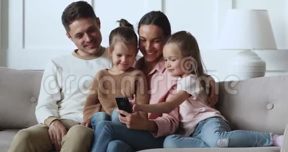 快乐的父母带着女儿在沙发上用智能手机放松视频的预览图