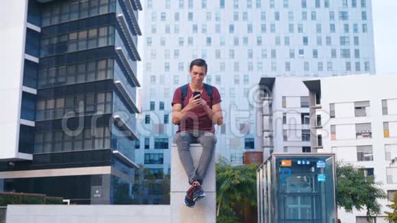 一个快乐的年轻人坐在商业区在现代玻璃的背景下使用他的智能手机视频的预览图