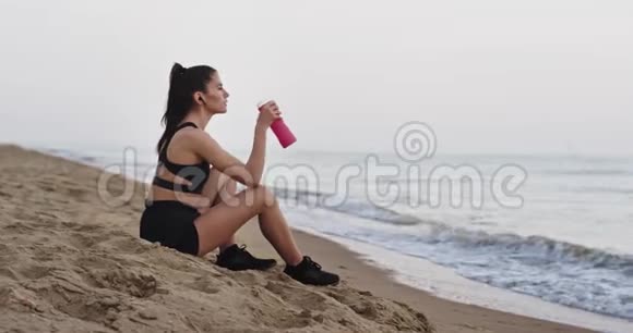 早上锻炼后女士在海边的海滩上休息喝着水享受着视频的预览图