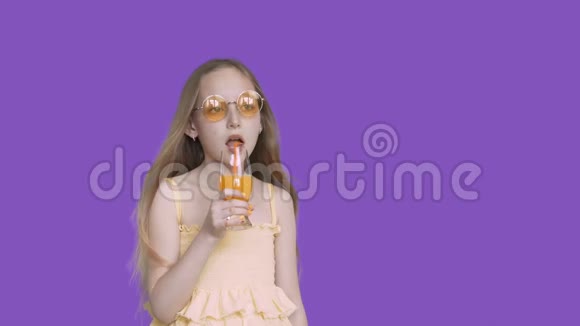 少女在紫罗兰色的背景下用吸管喝橙汁戴着黄色太阳镜喝酒的年轻女孩视频的预览图