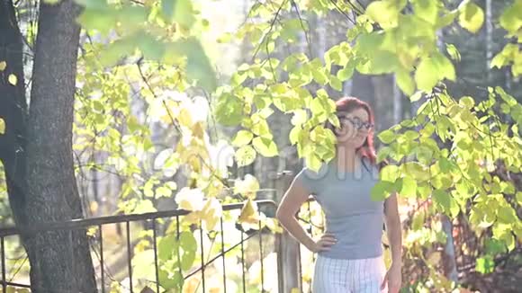 戴着眼镜的美丽悲伤的红发女子独自站在森林里穿着灰色T恤和条纹的女汉子视频的预览图