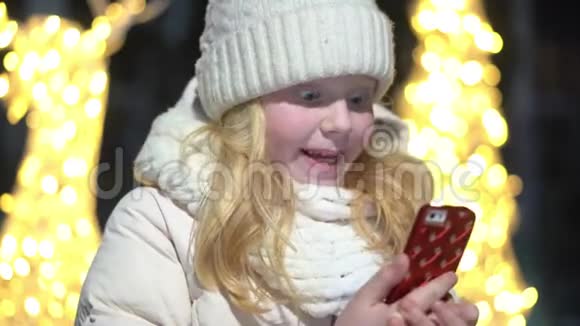 冬天一个漂亮的金发女孩在圣诞节装饰的街道上用智能手机聊天视频的预览图