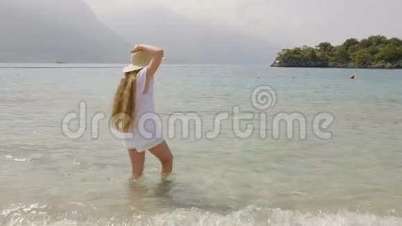 长头发的女孩戴着草帽穿着白色的衣服站在透明的海水中在山的背景上旅游女孩视频的预览图
