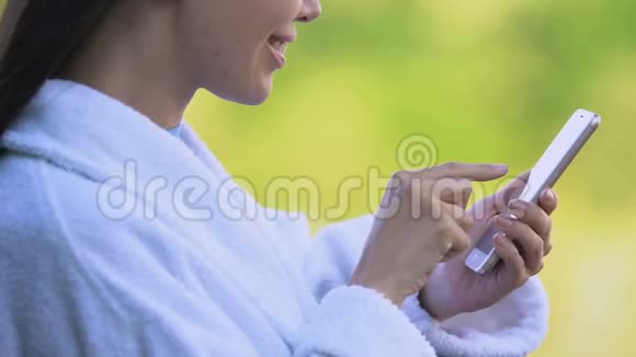 女士穿着浴袍在度假露台上滚动智能手机服务订购视频的预览图