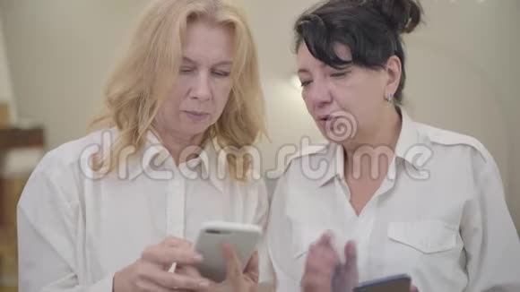 高加索族女性用智能手机教她的朋友两位穿白衬衫的成熟女士的肖像视频的预览图