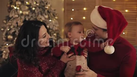 年轻微笑的白种人家庭和小男孩看着欢快的微笑坐在沙发上靠近圣诞树视频的预览图