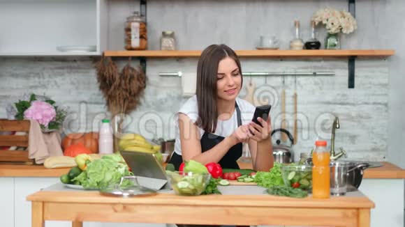 穿着围裙在厨房里用智能手机做饭聊天的漂亮女人视频的预览图